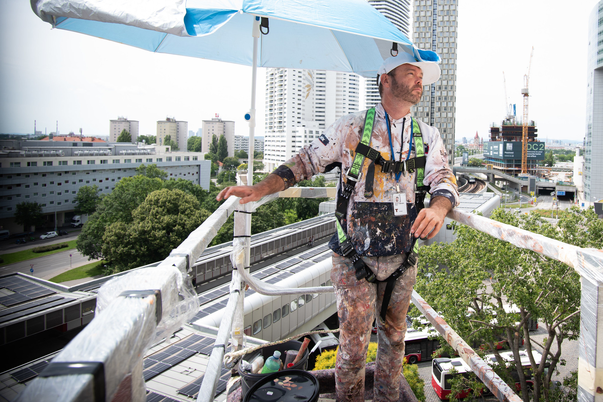 Man dressed in paint-covered clothes, wearing a cap and a safety harness, standing in a crane lift with blue sun umbrella in one corner, high up off the ground.